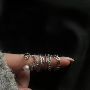 Sju silverringar med pärldetalj - Säljer ett set med sju snygga silverfärgade ringar i olika former och mönster. Några har tvinnad design, kedjelänk och hjärtan, en har en vit pärldetalj och en annan glittriga stenar. Perfekt för att mixa och matcha på flera fingrar.