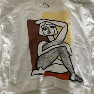 Vit sweatshirt med konstnärligt tryck - Säljer en vit sweatshirt med ett stort grafiskt tryck framtill i brunt, gult och orange. Motivet föreställer en stiliserad person i svart och vitt med coola linjer och blockfärger. Tröjan har rund hals och långa ärmar, perfekt för dig som gillar unik design.