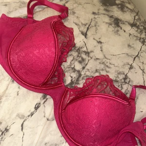 Rosa spets-bh med pushup och detaljer - Snygg rosa bh med vadderade kupor och spetsdetaljer längs kanterna. Axelbanden är justerbara och bh:n har en feminin design med blommönstrad spets framtill. Perfekt för dig som vill ha något extra under kläderna.