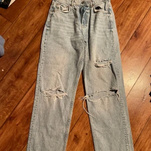 Ljusblå ripped jeans med raka ben - Säljer ett par ljusblå jeans med raka ben och slitna detaljer på båda benen. Klassisk femficksmodell med knapp och dragkedja i gylfen. Perfekta för en avslappnad och trendig look.