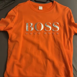 Orange tröja från Hugo Boss - Snygg orange tröja från Hugo Boss med stort silverfärgat BOSS-tryck på bröstet. Klassisk rund halsringning och korta ärmar. Perfekt för dig som vill sticka ut med en färgstark och trendig look.