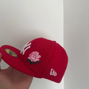 Röd New Era 59FIFTY keps med ros - Snygg röd New Era 59FIFTY keps med broderad vit NY-logga och en rosa ros med gröna blad på framsidan. På sidan finns ett broderat 1989-märke i grönt och vitt. Undersidan av skärmen är ljusrosa. Perfekt för dig som vill sticka ut med coola detaljer.