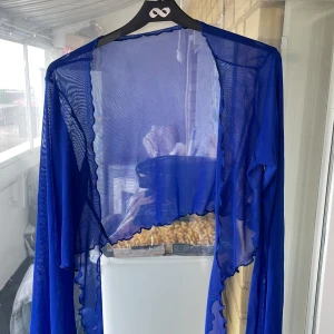 Blå transparent bolerotopp i mesh - Säljer en blå transparent bolerotopp i mesh med vågiga kanter. Toppen har lång ärm och är öppen framtill, perfekt att slänga över ett linne eller klänning för en cool layering-effekt. Materialet är lätt och luftigt. 