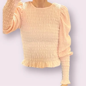 Ljusrosa smockad Blus med puffärm och ribbad - Supersöt ljusrosa blus med smockat tyg och puffiga långärmar. Toppen har en rund halsringning och volangkant nertill för extra detalj. Perfekt för dig som gillar romantisk och trendig stil.