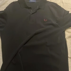 Svart pikétröja från Fred Perry - Klassisk svart pikétröja från Fred Perry. Tröjan har krage, knäppning framtill och den ikoniska Fred Perry-loggan broderad i rött på bröstet. Perfekt för en stilren och sportig look.