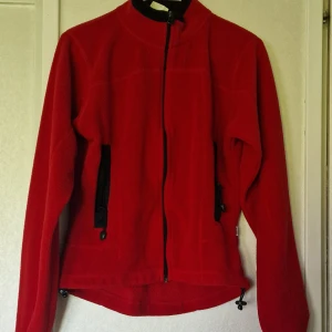 Röd fleecetröja från Topline - Röd fleecetröja från Topline med hel dragkedja framtill och svarta detaljer vid fickorna och kragen. Jackan har hög krage, långa ärmar och justerbar dragsko nedtill. Perfekt för kyliga dagar när du vill vara både bekväm och snygg.  Det är möjligt att den är något tvättpåverkad i färgen och det finns noppror.