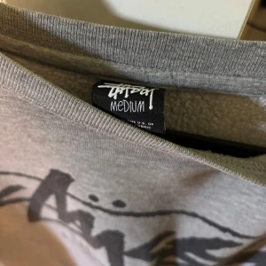 Grå långärmad Stüssy tröja med tryck - Säljer en grå långärmad tröja från Stüssy med stort svart tryck fram och bak. Tröjan har rund halsringning och är i mjuk bomullsmix, perfekt för streetwear-stil. Ikoniska städer tryckta på både fram- och baksidan.