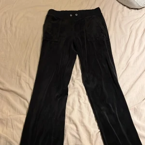 Svarta velourbyxor Juicy Couture  - Svarta mjukisbyxor från Juicy Couture i velour med bred resår i midjan, stora fickor fram och bak med knappar samt broderad JC-logga. Klassisk relaxed passform och snyggt glansigt material som ger en lyxig känsla. Storlek S