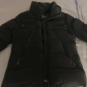 Svart pufferjacka från Moncler Grenoble - Säljer en svart pufferjacka från Moncler Grenoble med hög krage och flera dragkedjeförsedda fickor. Jackan är vadderad och har en clean, sportig look med diskreta logodetaljer. Perfekt för kalla dagar när du vill hålla stilen och värmen. 