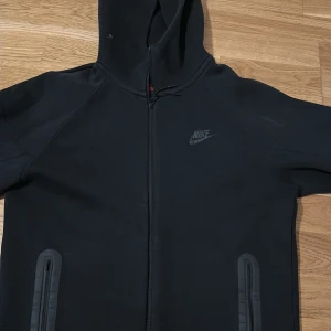 Svart Nike hoodie med dragkedja - Svart hoodie från Nike med hel dragkedja framtill och två fickor med dragkedja. Tröjan har huva och diskret Nike-logga på bröstet. Perfekt för en sportig och avslappnad stil.