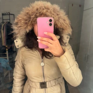 Beige Moncler pufferjacka med päls - Säljer en beige pufferjacka från Moncler med fluffig päls runt huvan och markerad midja med bälte. Jackan har dragkedja framtill och snygga detaljer som Moncler-logga på dragkedjan. Perfekt för kalla dagar när du vill vara både varm och stilren.