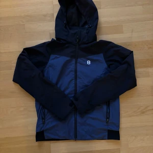 Blå och svart vindjacka från 8848 Altitude - Snygg vindjacka från 8848 Altitude i mörkblå och svart med huva och hel dragkedja framtill. Jackan har två sidofickor med dragkedja och en stilren 8-logga på bröstet. Perfekt för dig som gillar sportig och clean stil.