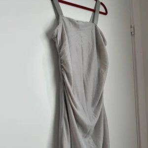 Lång silverglittrig klänning med drapering - Snygg långklänning i silver med glittrigt tyg och draperad detalj längs sidan. Klänningen har breda axelband och en elegant, figurnära passform som ger en lyxig känsla. Perfekt för dig som vill sticka ut med en stilren look.