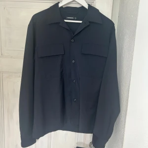 Svart overshirt från J.Lindeberg M - Stilren svart overshirt från J.Lindeberg i polyesterblandning. Skjortan har klassisk krage, två stora bröstfickor med lock och knäppning framtill. Observera att det är ett mycket litet hål på högra axeln! Förutom det lilla hålet är overshirten i mycket gott skick