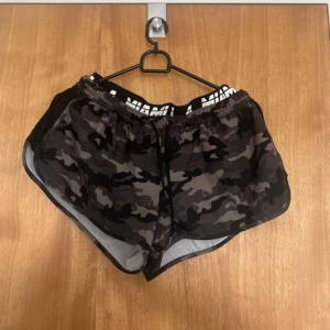 Censored camo shorts med snörning - Snygga camouflagemönstrade shorts från Censored i svart och grått. De har elastisk midja med snörning och vit logotyp på insidan av linningen. Perfekta för träning eller chill dagar. Lätta och luftiga i syntetmaterial.