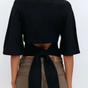 Svart croppad topp med knyt i ryggen - Snygg svart croppad topp med halvlånga ärmar och bred knytning i ryggen som ger en öppen rygg. Toppens stilrena design gör den lätt att matcha och den har en figurnära passform. Perfekt för dig som gillar trendiga detaljer och enkel stil.