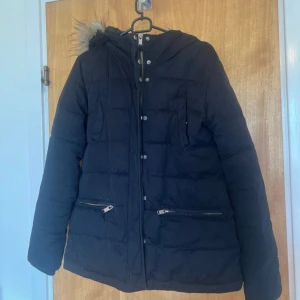 Svart pufferjacka från 365, strl 38 - Svart pufferjacka från 365 med huva och fuskpälsdetalj. Jackan har dragkedja och knappar framtill, två snedställda fickor med dragkedja och quiltad design. Perfekt för kalla dagar och riktigt skön att bära.