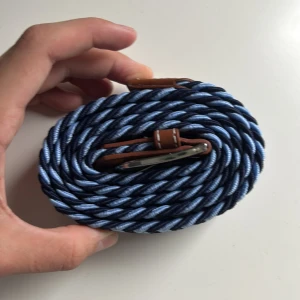 Blått flätat bälte med läderdetaljer - Snyggt flätat bälte i marinblått och ljusblått med bruna läderdetaljer och silvrigt spänne. Bältet har ett coolt mönster och passar perfekt till jeans eller chinos. Flexibelt material som gör det enkelt att justera längden.