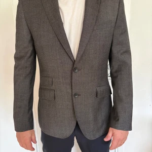 Grå kavaj från The Tailoring Club - Snygg grå kavaj från The Tailoring Club i ren ull. Klassisk modell med två knappar framtill, tre fickor med lock och slät finish. Fodrad insida i mörkblått tyg och diskreta sömdetaljer. Storlek 52. 