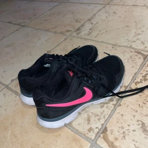 Svarta Nike sneakers med rosa swoosh - Snygga svarta Nike sneakers med rosa swoosh och detaljer. Skorna har vit sula, klassisk snörning och mesh-överdel för bra andningsförmåga. Perfekta för dig som gillar sportig stil och vill ha bekväma skor till vardagen. Lite slitna längs med hälen
