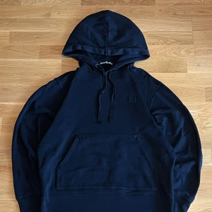 Svart Acne Studios Face Hoodie - Säljer en stilren svart hoodie från Acne Studios. Klassisk modell med huva, snörning och stor magficka framtill. Diskret Acne Face-logga på bröstet. Använd någon estaka gång och är i nyskick
