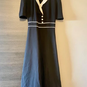 Svart långklänning med vit krage - Supersnygg svart långklänning med vit krage och vita knappar framtill. Klänningen har korta ärmar, markerad midja med vita ränder och en klassisk v-ringning. Materialet känns mjukt och har ett fint fall. Perfekt för dig som gillar retrovibbar och stilrena detaljer.