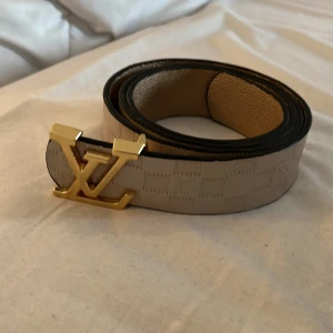 Beige Louis Vuitton skärp med guldspänne - Snyggt beige Louis Vuitton bälte med klassiskt rutigt mönster och ett stort LV-spänne i guld. Bältet är gjort i exklusivt skinn och har svarta kanter. Perfekt accessoar för att lyfta din outfit med lyxig vibe.
