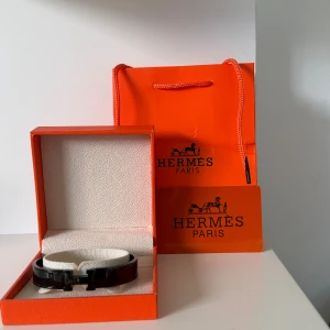 Svart armband från H - Säljer ett stilrent svart armband från H med klassisk H-detalj framtill. Armbandet kommer i original orange ask och påse, perfekt för dig som gillar lyxiga accessoarer med ikonisk design.