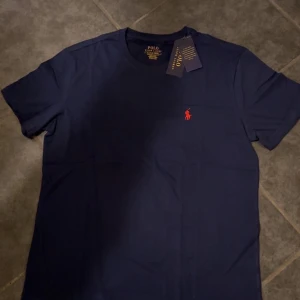 Mörkblå t-shirt från Polo Ralph Lauren - Snygg mörkblå t-shirt från Polo Ralph Lauren med klassisk röd logga broderad på bröstet. T-shirten har rund halsringning och korta ärmar. Tillverkad i mjuk bomull som känns skön mot huden. Perfekt för en clean och stilren look.
