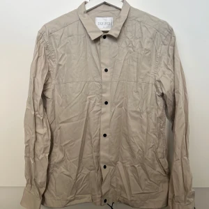 Beige skjorta - En beige skjorta med svarta knappar. Inte så använd men i fotot är den tyvärr inte struken. 