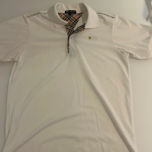 Vit Burberry piké - Vit pikétröja från Burberry i storlek L med klassisk krage och korta ärmar. Ikoniskt rutigt mönster på insidan av kragen och knappslån. Broderad Burberry-logga och emblem på bröstet. Tillverkad i bomull för en skön och stilren känsla.
