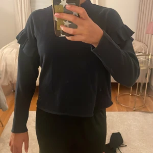 Svart långärmad tröja med volang - Svart långärmad tröja med volangdetaljer på axlarna och rak passform. Tröjan har en högre rund halsringning och är tillverkad i ett mjukt material som känns skönt mot huden. Perfekt för dig som gillar stilrena plagg med en twist.