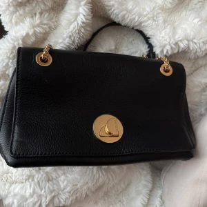 Svart axelväska från Coccinelle i skinn - Snygg svart axelväska från Coccinelle i äkta italienskt skinn. Väskan har guldfärgade kedjedetaljer och ett elegant, runt spänne framtill. Stilren och klassisk design med exklusiv känsla, perfekt för dig som gillar lyxiga accessoarer.