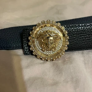  svart skärp med Medusa-spänne - Svart skärp i präglat skinn med ett stort, runt guldspänne dekorerat med gnistrande stenar och den ikoniska Medusa-loggan. Bältet har en lyxig och iögonfallande design som verkligen sticker ut.