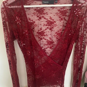 Vinröd spetsblus med paljetter  - Säljer en vinröd långärmad spetsblus från KappAhl med djup v-ringning och omlottkänsla. Blusen har blommigt spetsmönster och är dekorerad med små paljetter som ger extra glans. Perfekt för dig som vill sticka ut lite extra.