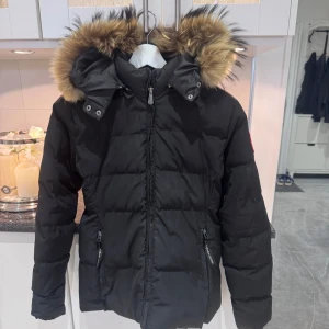 Svart dunjacka från Canada Goose - Säljer en svart dunjacka från Canada Goose med fluffig pälskant på huvan. Jackan har dragkedja framtill, två fickor med dragkedja och klassisk patch på ärmen. Perfekt för kalla vinterdagar och riktigt snygg streetstil. Storlek S