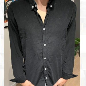 Svart långärmad skjorta med knappar - Svart skjorta med klassisk krage och vita knappar hela vägen framtill. Skjortan har lång ärm och en avslappnad passform, perfekt för en stilren look. Tillverkad i mjuk bomull som känns skön mot huden.