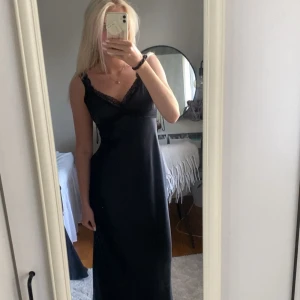 Svart långklänning med spetsdetalj - Säljer en svart långklänning med smala axelband och v-ringning. Klänningen har fina spetsdetaljer vid urringningen, klänningen är endast testad med prislappen kvar. Har tyvärr inte kommit till andväning än