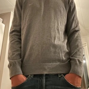 Grå långärmad tröja i bomull - En stilren grå långärmad tröja i mjuk bomull. Tröjan har en klassisk passform med ribbade muddar vid ärmslut och nederkant. Perfekt för lager-på-lager och enkel att matcha med jeans eller andra byxor.