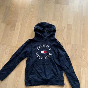 Marinblå hoodie från Tommy Hilfiger - Snygg marinblå hoodie från Tommy Hilfiger med klassisk logga och broderad text framtill. Tröjan har huva, känguruficka och långa ärmar. Perfekt för dig som gillar streetwear och vill ha en stilren look.