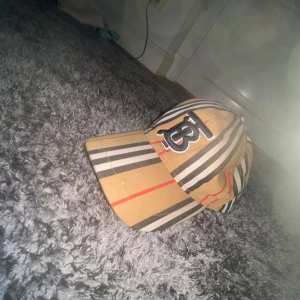 Burberry randig keps med logga - Snygg Burberry keps i klassiskt beige med svarta, vita och röda ränder. Framtill sitter en stor broderad TB-logga i svart och vitt. Kepsen är tillverkad i bomull och har justerbar rem baktill. Perfekt för dig som vill sticka ut med en ikonisk accessoar.