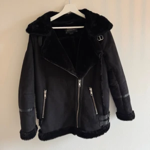 Svart biker pälsjacka från Pull&Bear - Snygg svart bikerjacka pälsjacka från Pull&Bear med fuskpälsfoder, stora slag och coola spänndetaljer vid krage och nederkant. Jackan har dragkedjor framtill och vid fickor samt sköna muddar i fuskpäls. Perfekt för kalla dagar och ger en edgy vibe