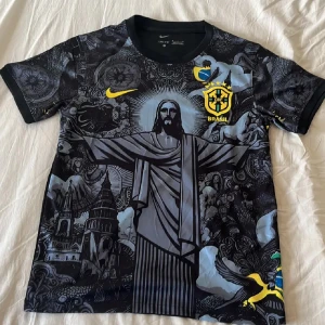 Nike Brasil Dri-Fit fotbollströja M - Unik svart Nike Brasil fotbollströja i Dri-Fit-material med detaljerat grafiskt tryck av Kristusstatyn och brasilianska motiv. Gula och gröna accenter, CBF-emblem på bröstet och korta ärmar. Perfekt för dig som vill sticka ut på planen eller läktaren.