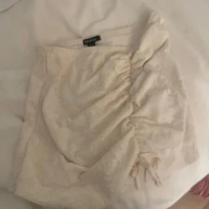 Beige kortkjol med snörning från Boohoo - Supersöt beige kortkjol från Boohoo med diskret blommönster och snygg snörningsdetalj framtill. Kjolen har en lätt volang och är perfekt för dig som gillar en feminin och trendig stil.