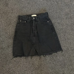 Svart jeanskjol  - Svart jeanskjol som är hög midja, knapp och dragkedja framtill samt två stora bakfickor. 