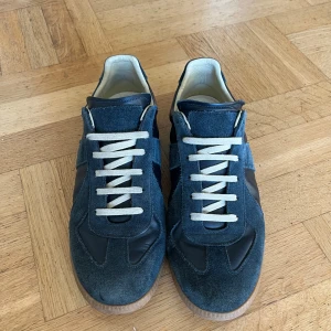 Blå sneakers från Maison Margiela - Kontakta vid prisförslag. Box och dustbag ingår!Maison Margiela sneakers i mörkblå mocka och skinn med vita snören och gummisula. Klassisk rund tå och platt sula, snygg kontrast mellan mocka och läder. Kommer med originalkartong och dustbag. Perfekt för dig som gillar stilrena och unika sneakers. Skorna har dock en skada där bak. Skorna är i storlek 43 men passar även 42