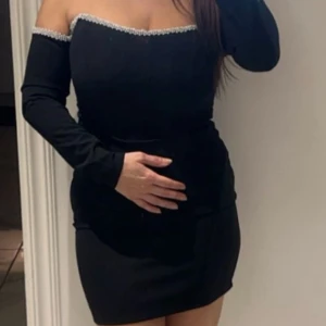 Svart offshoulder kortklänning - Snygg svart kortklänning med offshoulder och långa ärmar. Klänningen har en pärlprydd kant upptill som ger en extra touch. Figurnära passform som framhäver siluetten. Perfekt för dig som vill sticka ut med stilren elegans.