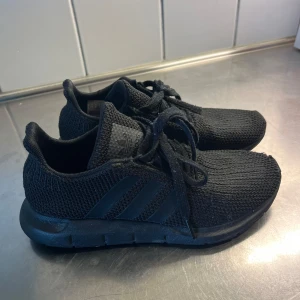 Svarta Adidas sneakers  - Säljer ett par helsvarta Adidas sneakers i storlek 38. Skorna har en stickad ovandel i mesh, klassiska tre ränder på sidan och snörning. Sulan är svart och ger en sportig vibe. Perfekta för dig som gillar stilrena och bekväma skor till vardags. ( skav vid tån ) 