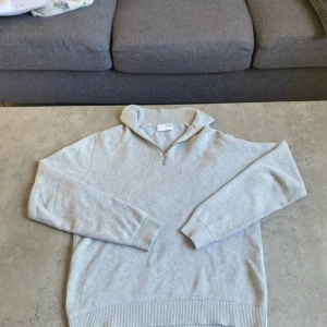 Ljusgrå half zip-tröja Selected Homme - Ljusgrå stickad tröja från Selected Homme med half zip och hög krage. Tröjan har ribbade muddar och nederkant, samt en klassisk passform. Perfekt för lager-på-lager och chill stil.