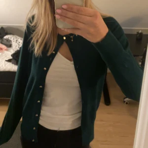 Mörkgrön kofta med guldknappar - Snygg mörkgrön kofta med långa ärmar och stilrena guldknappar framtill. Koftan har en klassisk passform och är tillverkad i ett mjukt stickat material som känns skönt mot huden. Perfekt att slänga över en t-shirt för en chill look.